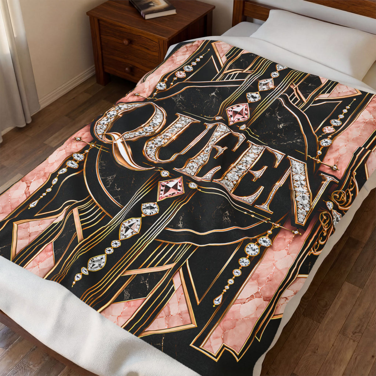A bedding set on a bed in a bedroom, featuring a throw blanket with a black and pink marble background, gold Art Deco lines, and the word 'QUEEN' in large jeweled letters, styled with white pillows and a wooden bedside table.