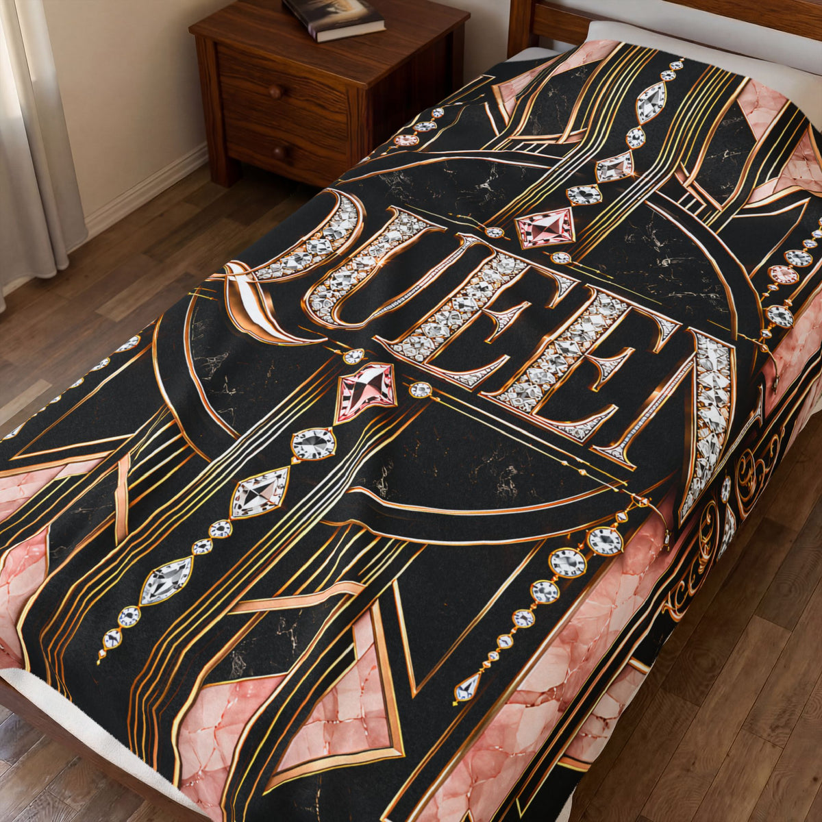 A bed dressed with a bold Art Deco-style throw blanket featuring the word 'QUEEN' in large jeweled letters over a black background with gold lines and pink marble accents, shown in a bedroom setting.