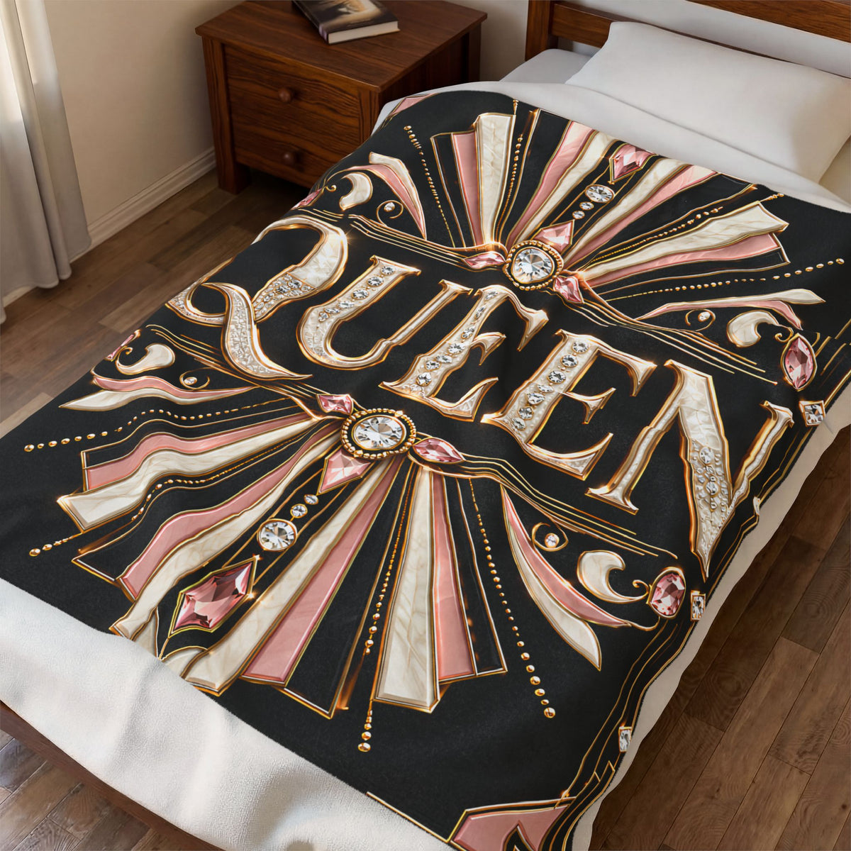 A black bedding blanket featuring a bold, ornate 'QUEEN' graphic in gold, pink, and cream with jewel-like accents, styled on a bed in a bedroom setting.