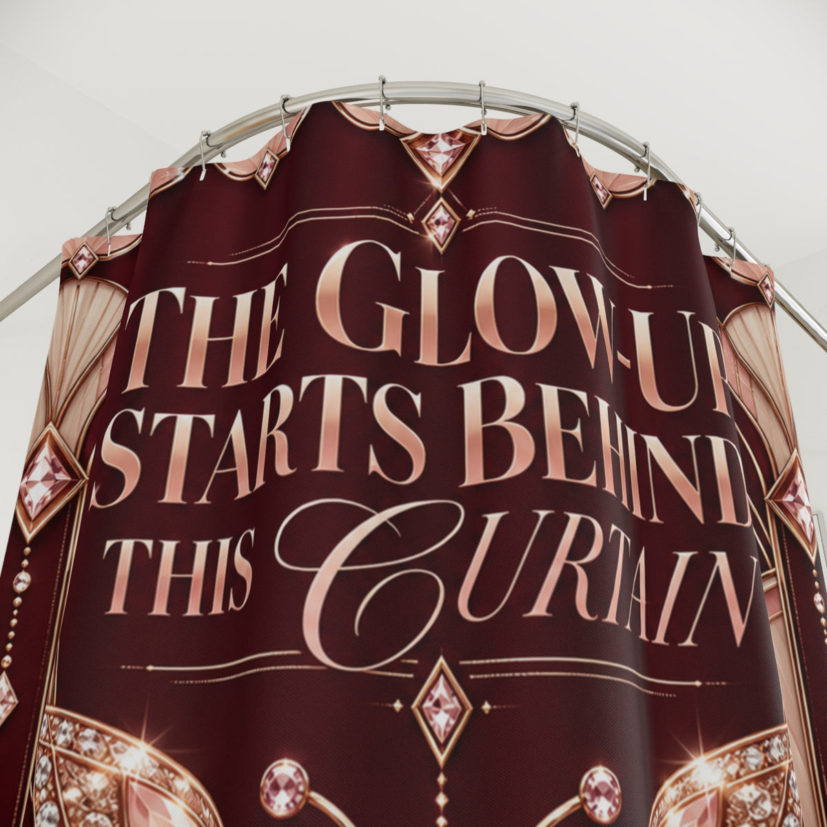 A maroon curtain with rose gold text reading 'THE GLOW-UP STARTS BEHIND THIS CURAIN' and decorative gem motifs, hanging from a metal rod against a light gray background.