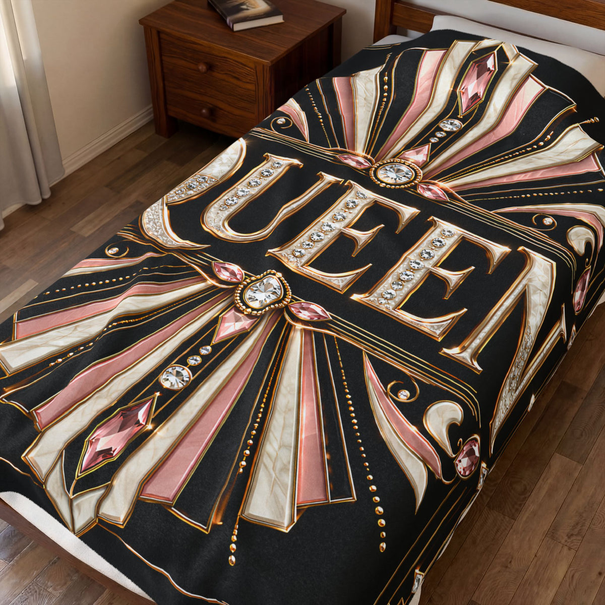 A bed in a bedroom styled with a black comforter featuring a large gothic-style 'QUEEN' design with pink, cream, and gold ornamental rays and jewel motifs. A wooden nightstand is visible in the background.