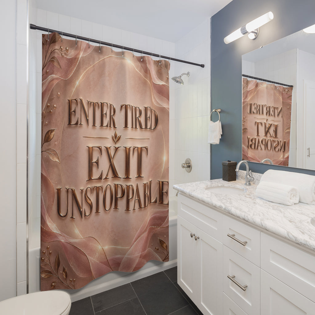 A bathroom scene showing a shower curtain with a printed design that reads 'ENTER TIRED EXIT UNSTOPPABLE' in large metallic-style lettering over a pink and rose-gold abstract background. The curtain is hanging in a modern bathroom with white cabinets, a marble countertop, a mirror, and dark gray floor tiles.