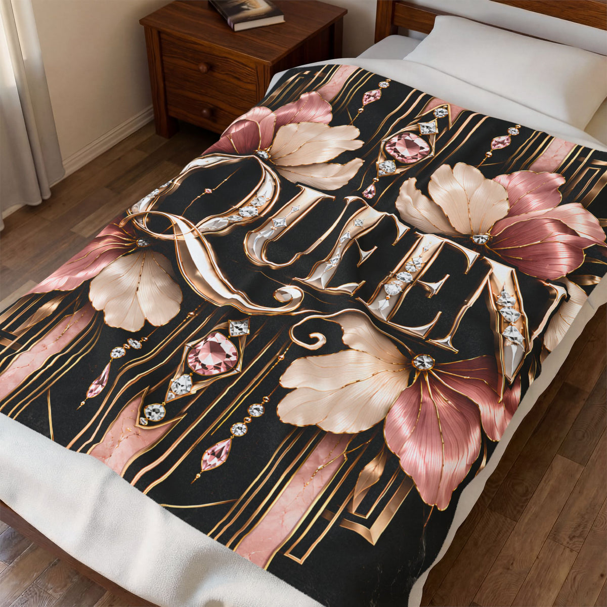A bed in a bedroom styled with a decorative printed blanket or comforter. The design features large pink and beige flowers, gold ornamental lettering, and pink gemstone motifs on a black background.