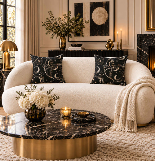 A luxurious cream bouclé sofa in a styled living room, decorated with two black throw pillows featuring a silver moon and star pattern, a cream knit throw blanket, and a round black marble coffee table with a gold base.