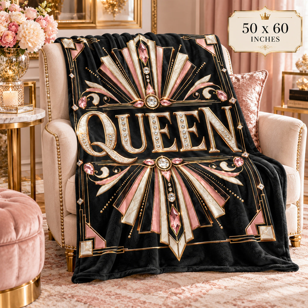 A plush black throw blanket draped over a cream armchair in a luxurious room setting. The blanket features a bold Art Deco design with the word 'QUEEN' in large gold letters, surrounded by pink and gold rays with faux jewel details.